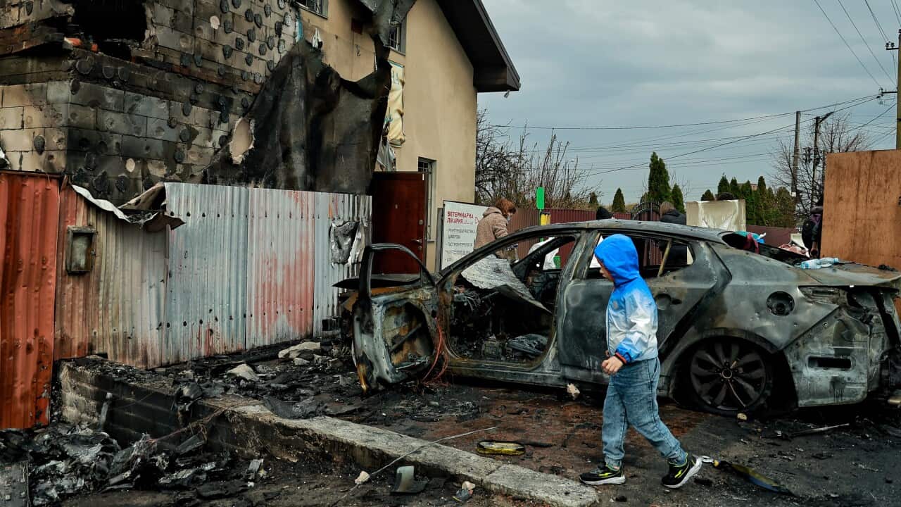 A young child in a blue hooded jacket walks past a burned-out car in front of a heavily damaged building.