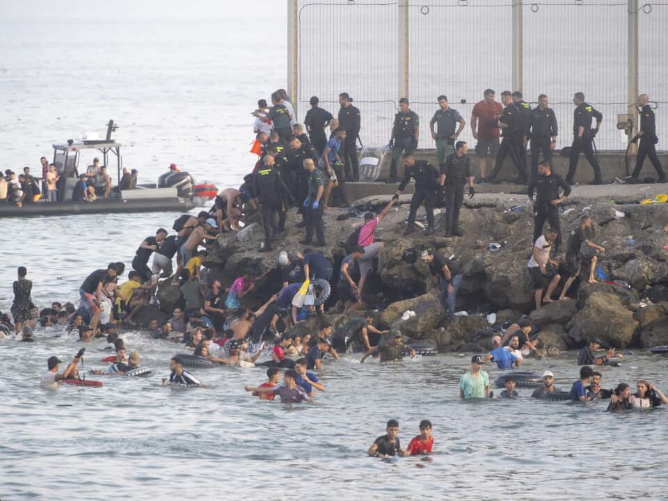 Dozens of migrants in the sea next to a rock breakwall. Uniformed police are standing on the breakwall, and a small boat carrying more migrants is in the background.
