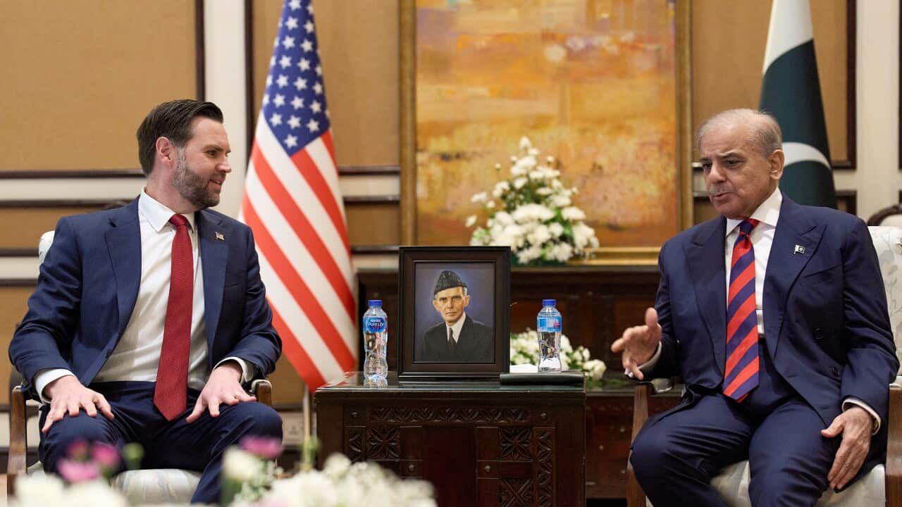 A younger white man and an older Pakistani man dressed in suits. They are seated and talking, an United States and Pakistan flag are behind them.