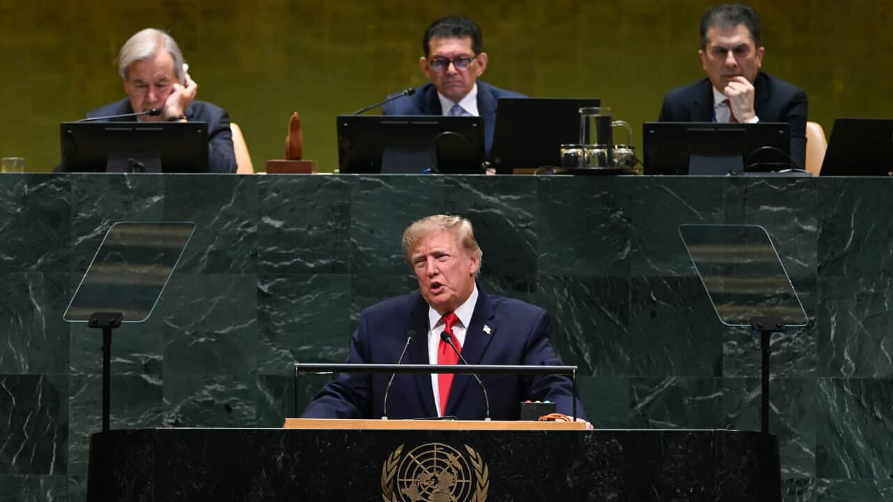 Man in a dark suit and red tie speaks from the United Nations General Assembly podium, with officials seated behind him and teleprompters on either side.