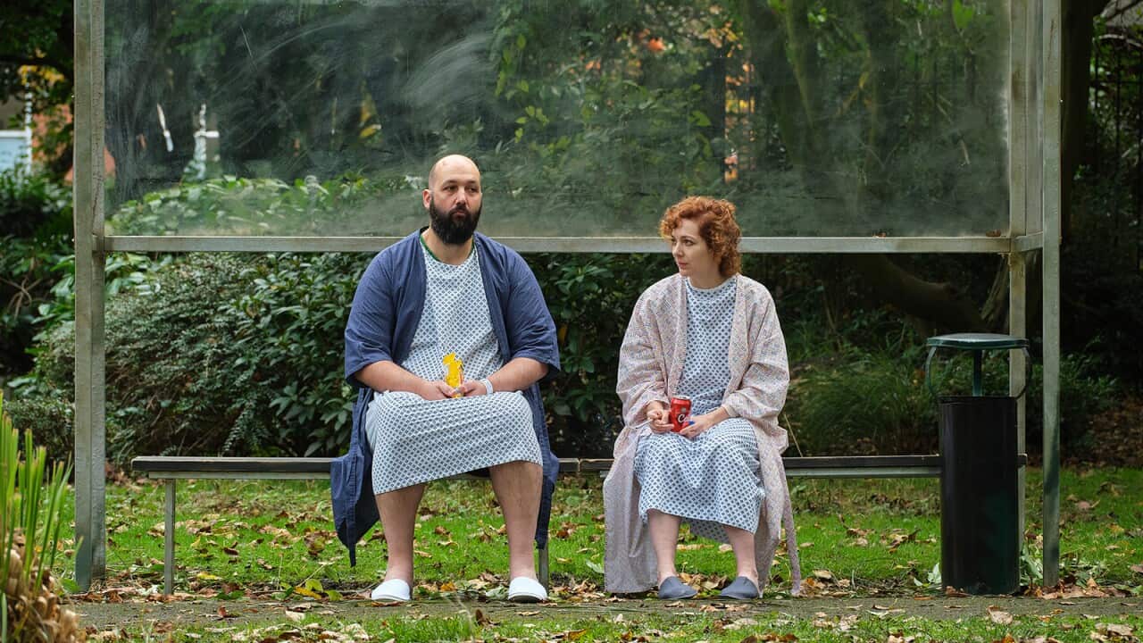 A man and a woman sit on a bench in a garden, both are dressed in hospital gowns. She looks sideways at him.