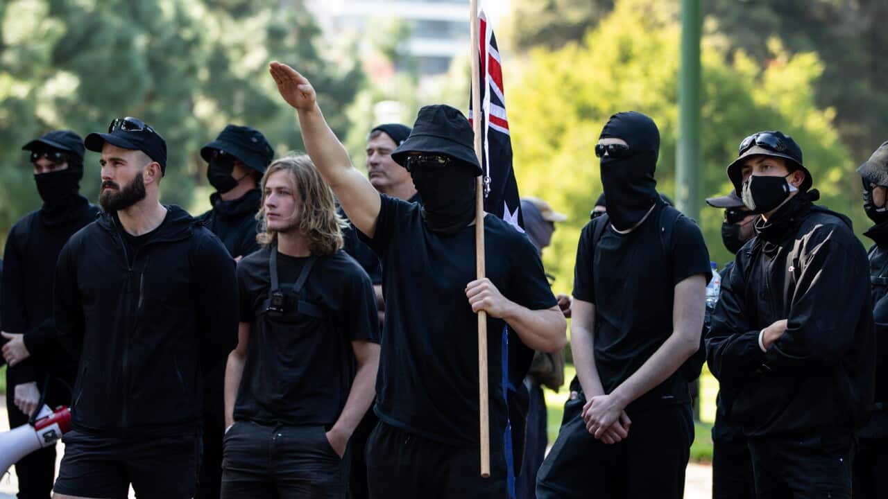 A small group of men standing outside and dressed in black, some wearing masks. One is holding an Australian flag on a pole and displaying a Nazi salute