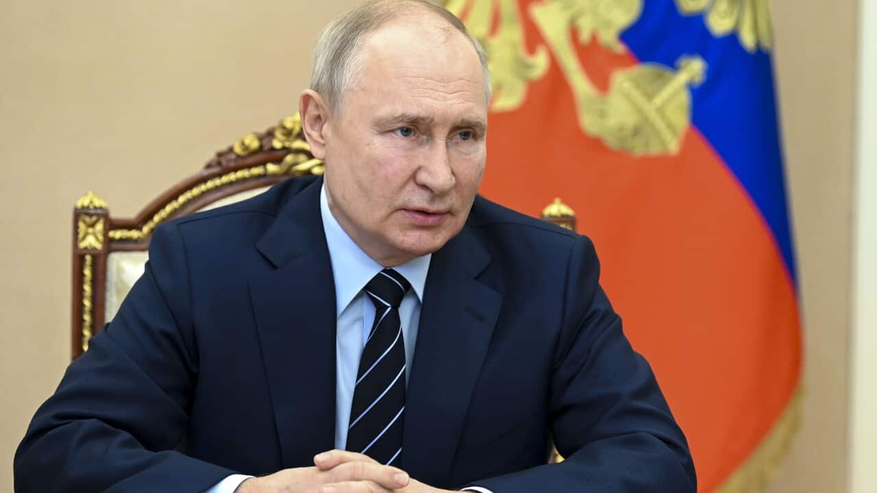 Vladimir Putin wearing a dark suit and sitting at a table. He has his hands clasped in front of him. A Russian flag is in the background.