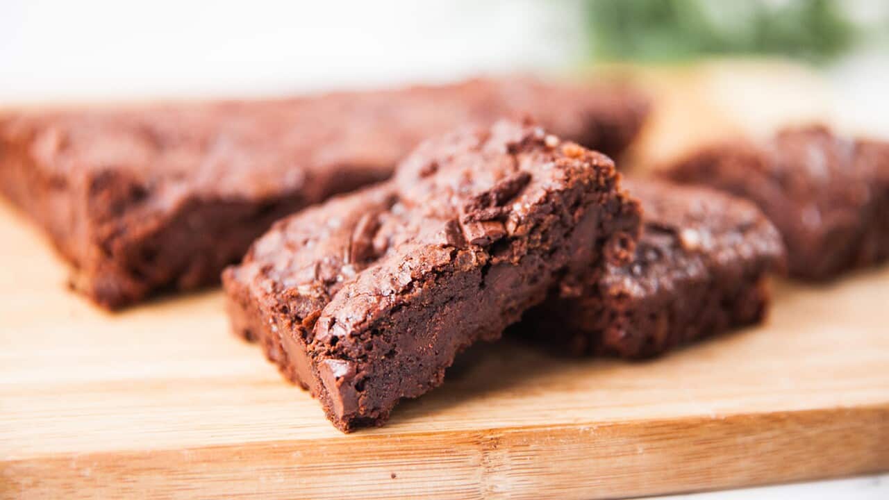 A fudgy piece of brownie sits on a wooden board. Behind it, two more pieces and a large uncut slab of brownie can be seen, out of focus.
