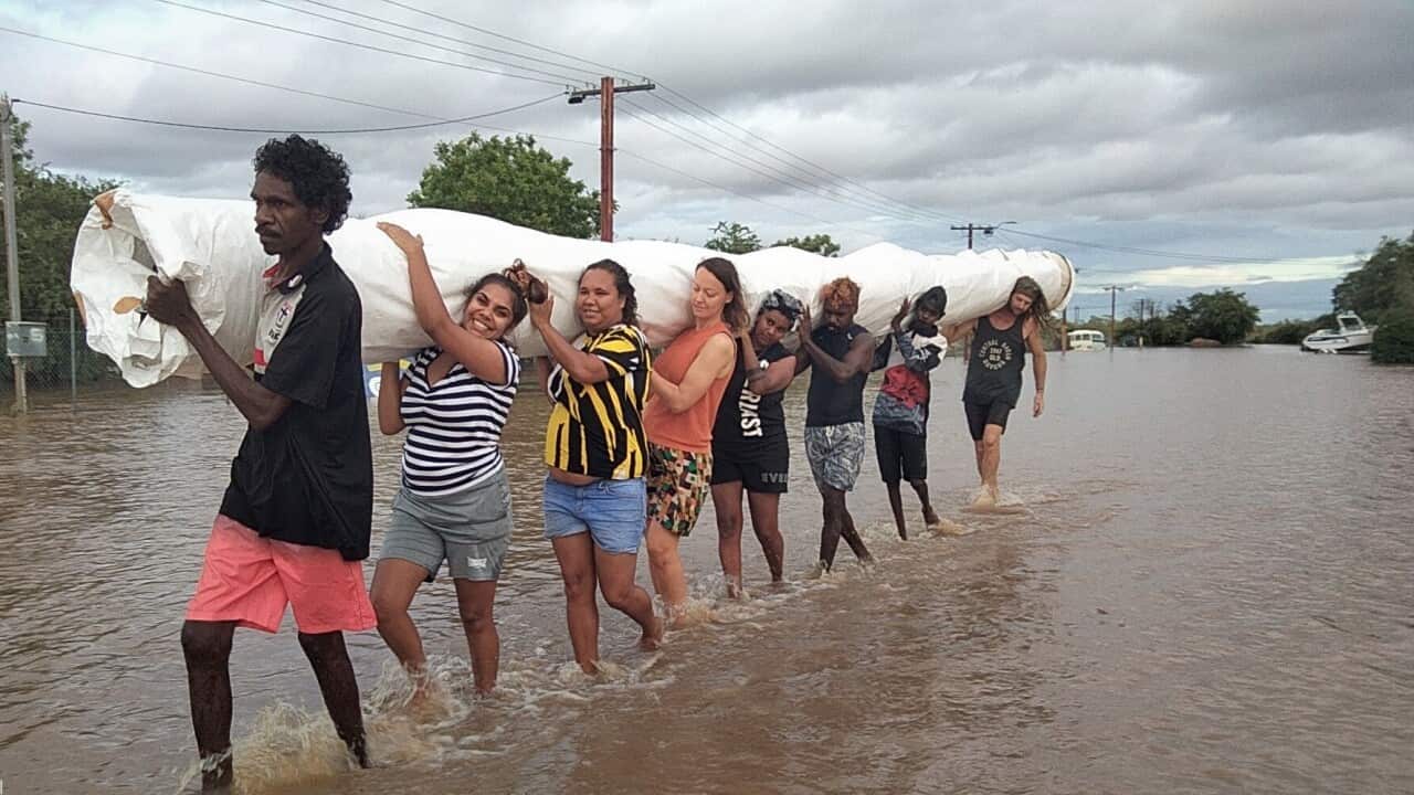 Eight people walk through ankle high water, each holding a part of a rolled up oversized canvas on their shoulder.