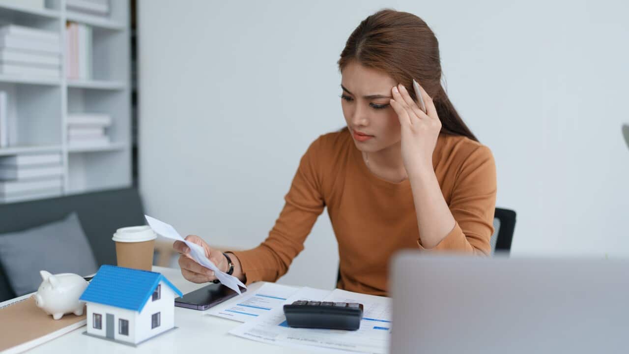 A stressed-looking woman sits at a table with sheets of paper, a laptop, a piggy bank and a model house in front of her.