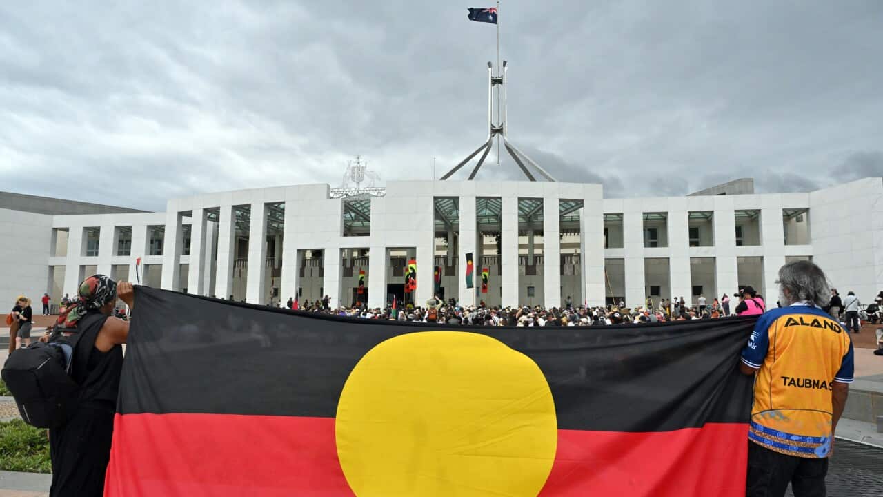 Two people hold up a large Aboriginal flag in front of Parliament House in Canberra. A crowd is gathered in front of them and the sky is grey.
