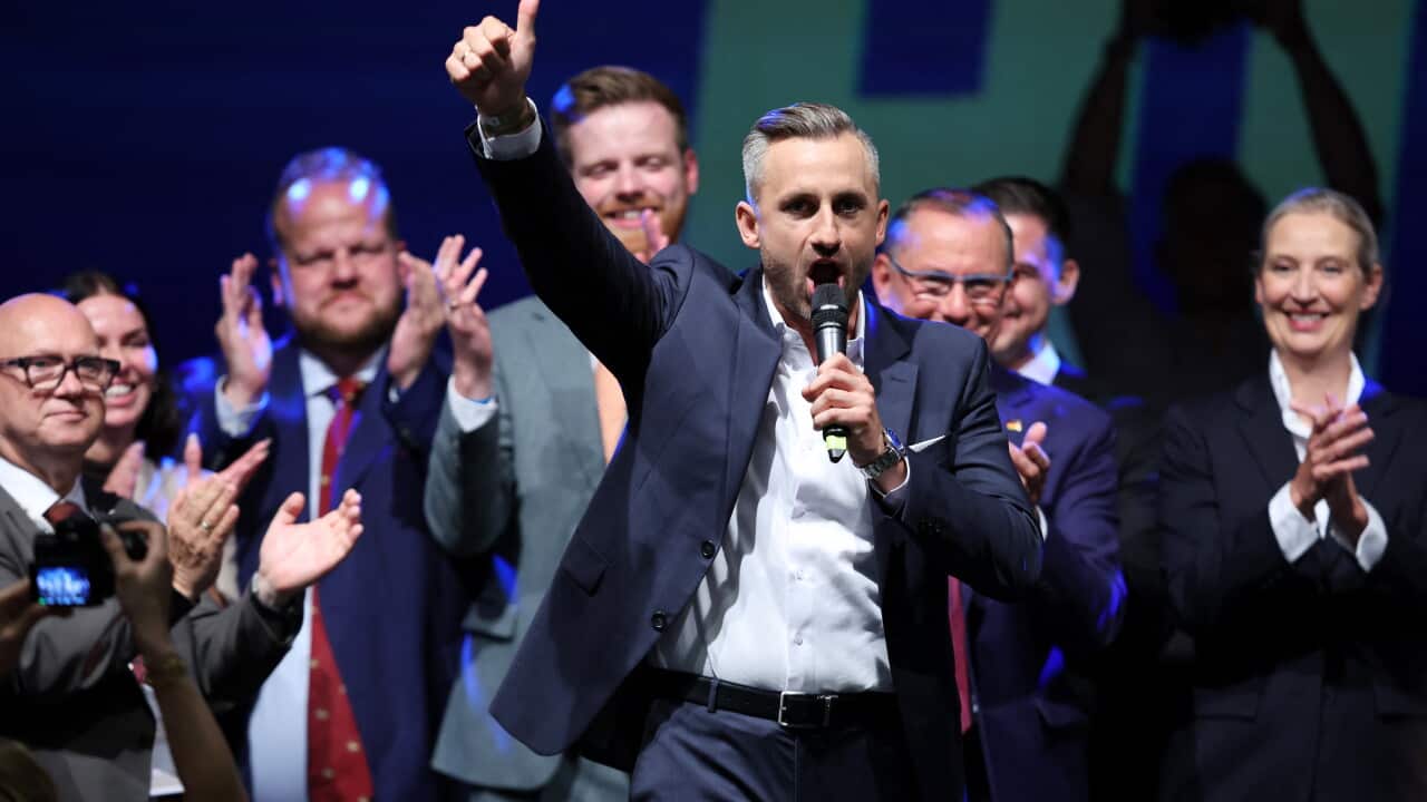 A man in a suit holds a microphone and gestures to the camera.