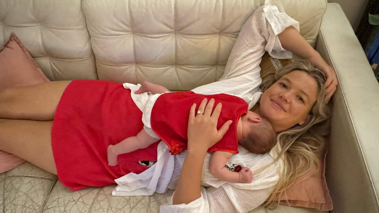 A young blonde white woman lying on a lounge and smiling. She is holding a baby in her arms.