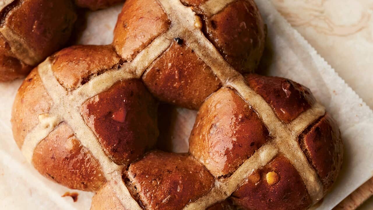 An angle six-pack of hot cross buns are seen from overhead, sitting on a sheet of baking paper. They glisten with a light glaze.