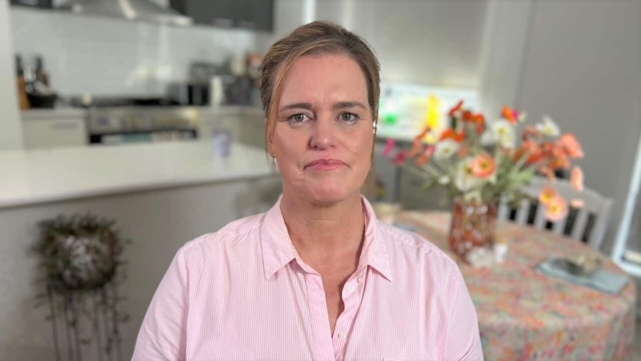 A woman with brown hair wearing a pink shirt sits in a kitchen in front of a bowl of flowers on a table.