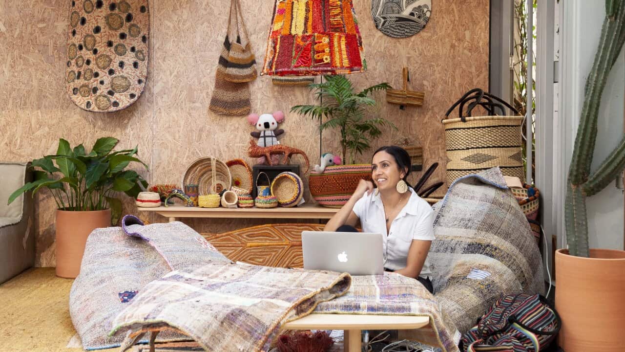 a WOMAN SITS ON THE the ground surrounded by woven fabrics, baskets, bags and more, with an open laptop before her