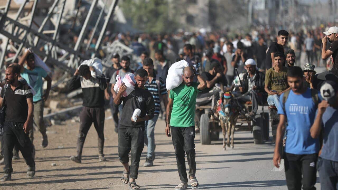 Crowd of displaced people carrying belongings through a damaged area.