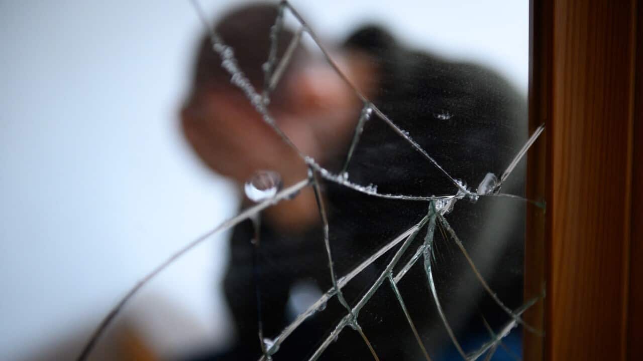 A man sits next to a cracked mirror on a bed and holds his head in his hands