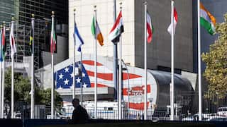 Flags from multiple countries line a plaza outside a large building featuring a mural of the US flag and the text “Freedom 250,” with a silhouetted pedestrian in the foreground.