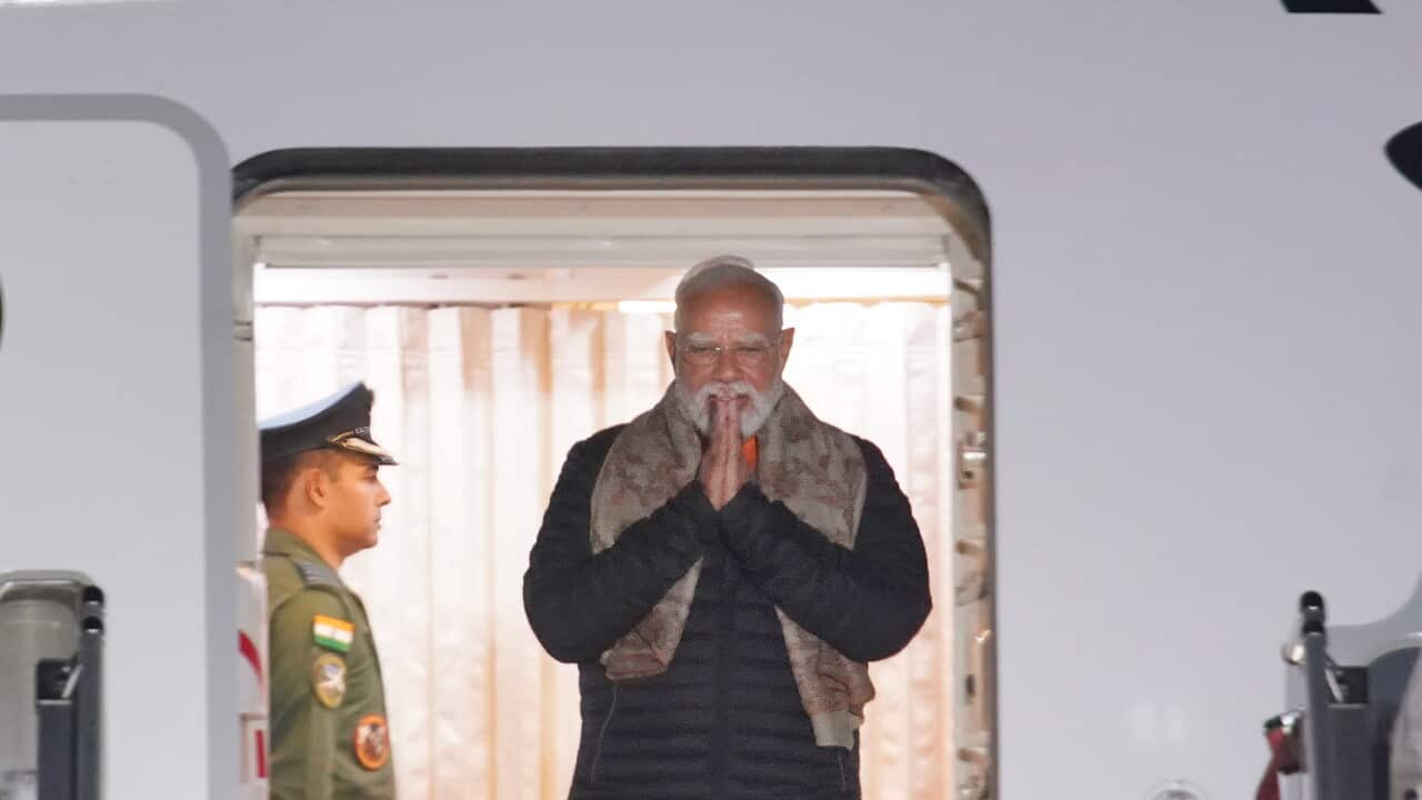 Narendra Modi, wearing a black top long-sleeved parka top and a brown scarf over his shoulders stands in the doorway of an aeroplane and presses his hands together near his mouth in greeting.