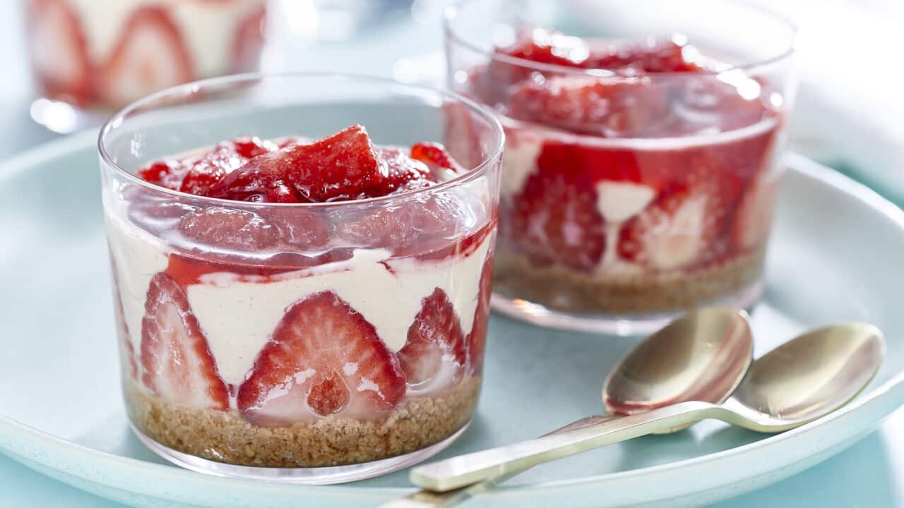 Two dessert serving glasses sit on a wide pale blue plate, along with two spoons. Each glass has a dessert with a crumb base later topped with strawberries and a pale mousse.