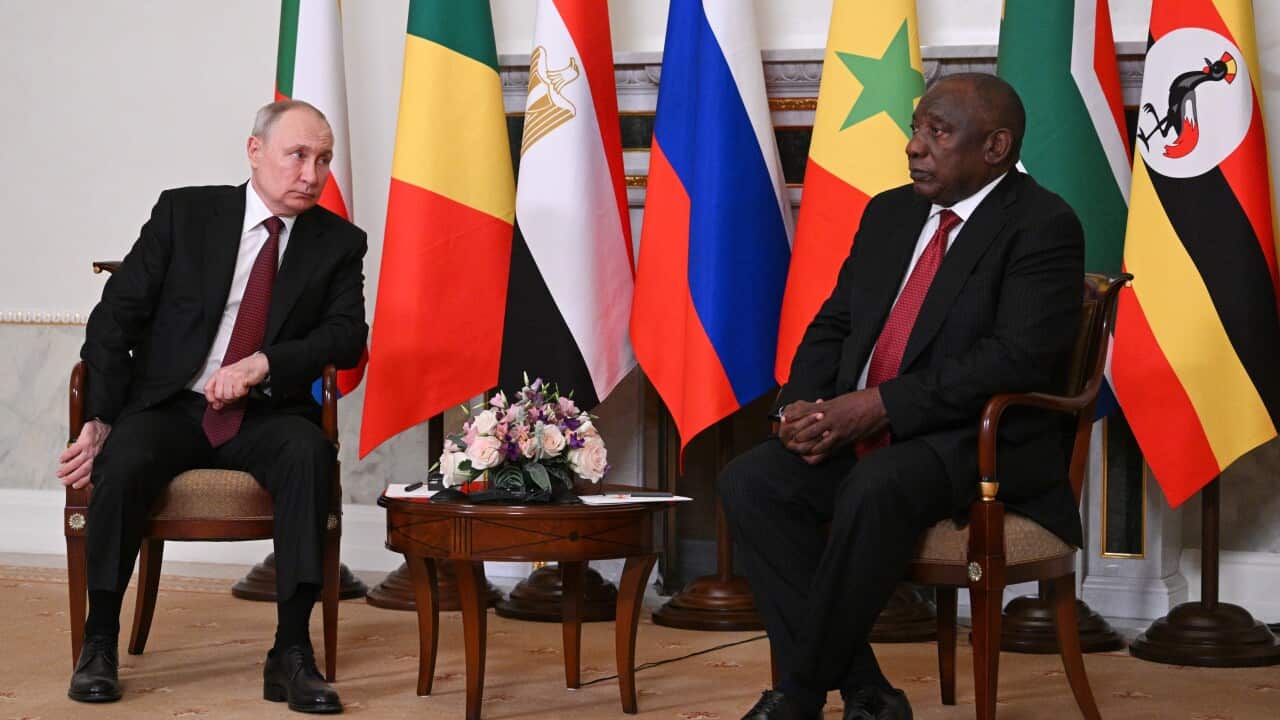 An older white man in a black suit seated next to a older black man in a suit. National flags hang on poles behind them.