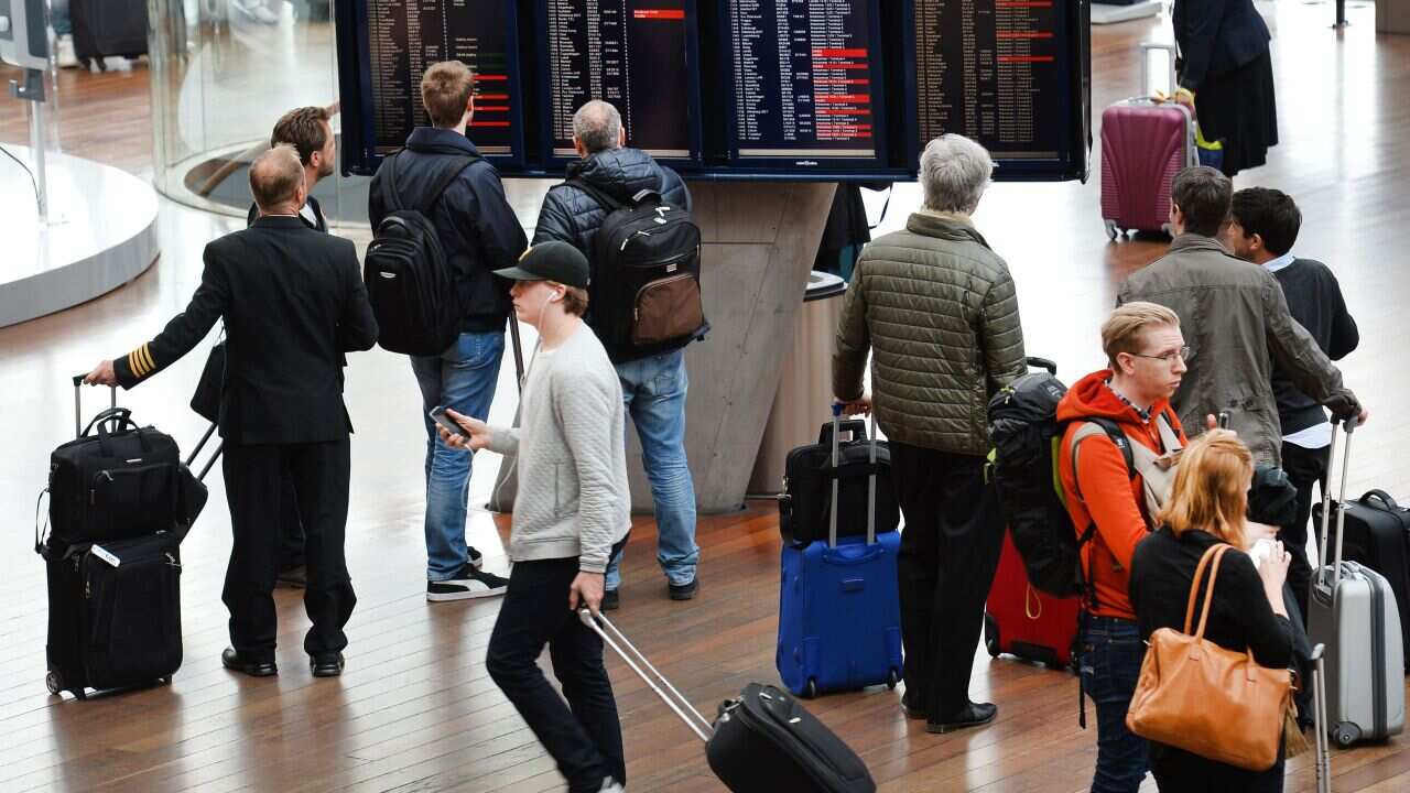 People in an airport terminal. Some are wheeling suitcases, others are looking at the arrivals and departures board.