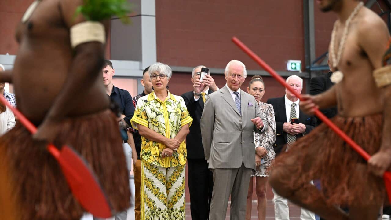 Two men in traditional dress being watched by a man in a grey suit and woman in colourful print suit