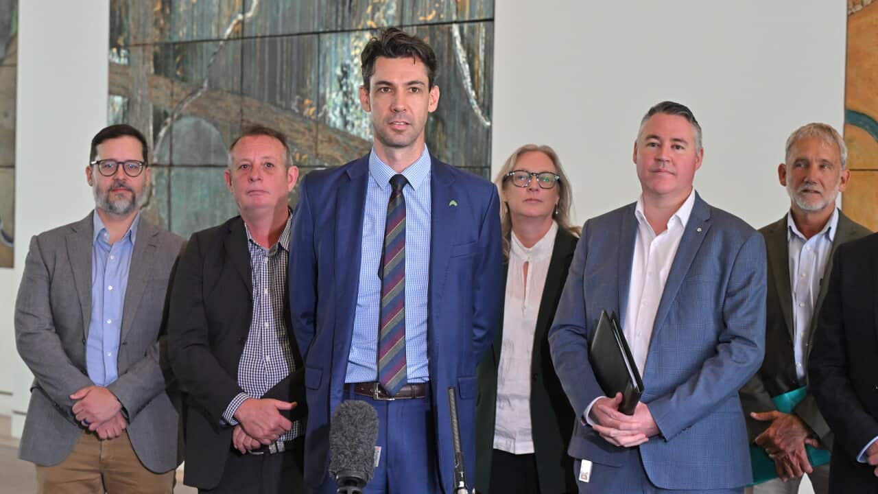 A tall man in a blue suit and striped tie stands at a microphone speaking to reporters, flanked by five other people in professional attire.