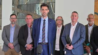 A tall man in a blue suit and striped tie stands at a microphone speaking to reporters, flanked by five other people in professional attire.