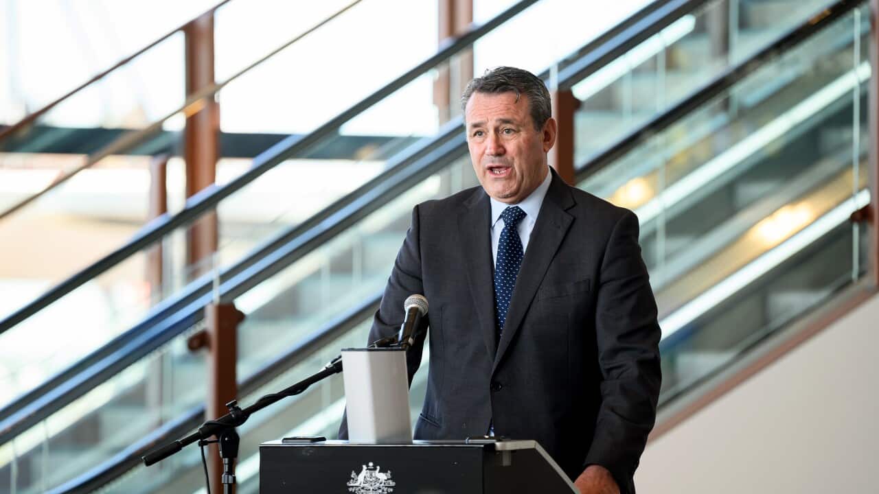 A man wearing a dark suit and blue tie speaks from a lectern bearing the Australian Government crest in front of a modern staircase.