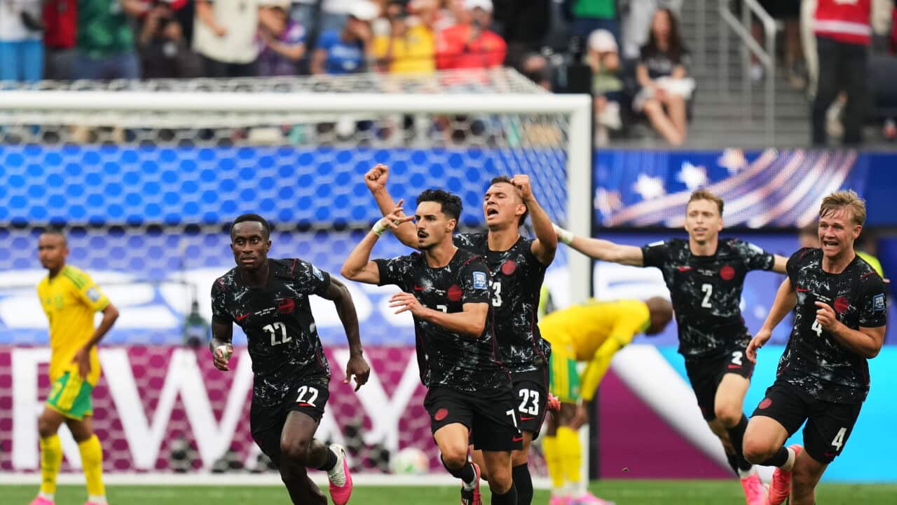 A football player in a black jersey celebrates a goal by making a gesture with his hand by his head. Four other players in black jerseys are running behind him elated as they celebrate. Two players in yellow and green jerseys are seen dejected in the background in front of the goalpost.