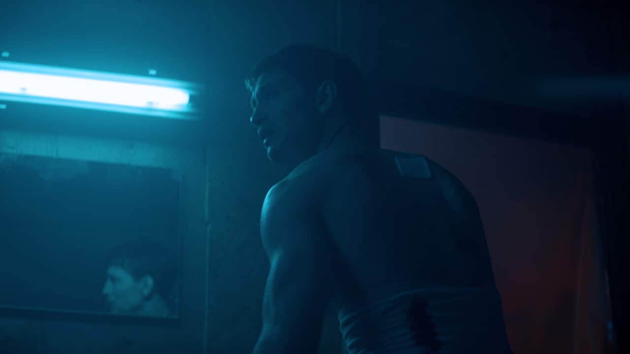 A man with a blood-soacked bandage around his waist and another bandage on his shoulder stands in front of a bathroom mirror. The light is dark and blue-tinged, and he looks to one side with a worried expression.