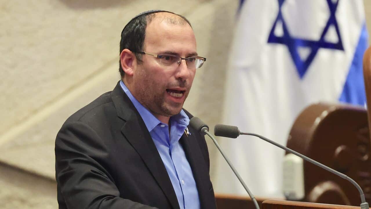 A man in a suit speaks from behind a lectern inside. There is an Israeli flag in the near distance