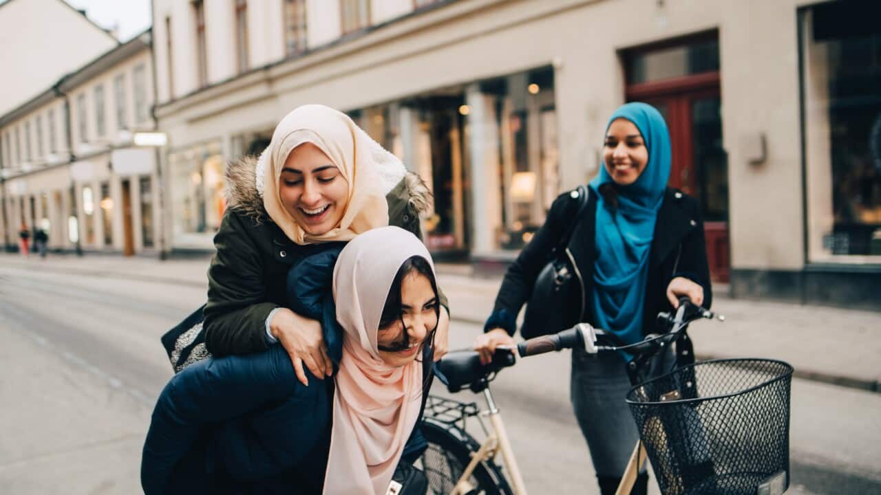 Cheerful teenage girl giving young woman piggyback by friend walking with bicycle on street in city