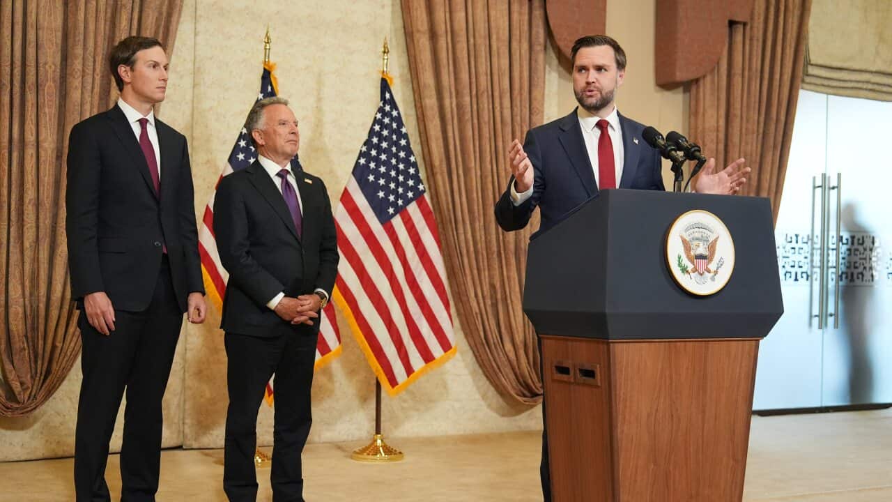 A white man in a suit, with brown hair and a beard, speaking at a podium. Two other men stand to his side, American flags hang on a flagpole.