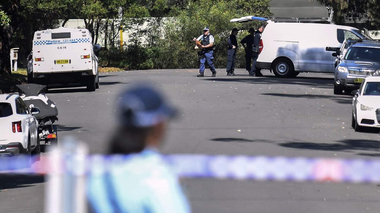 An out-of-focus police officer and police tape in the foreground, with police vans and officers in the background.