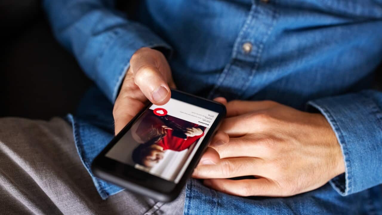 A close up of a man in a blue shirt on his phone, which has an image of a woman's dating profile.