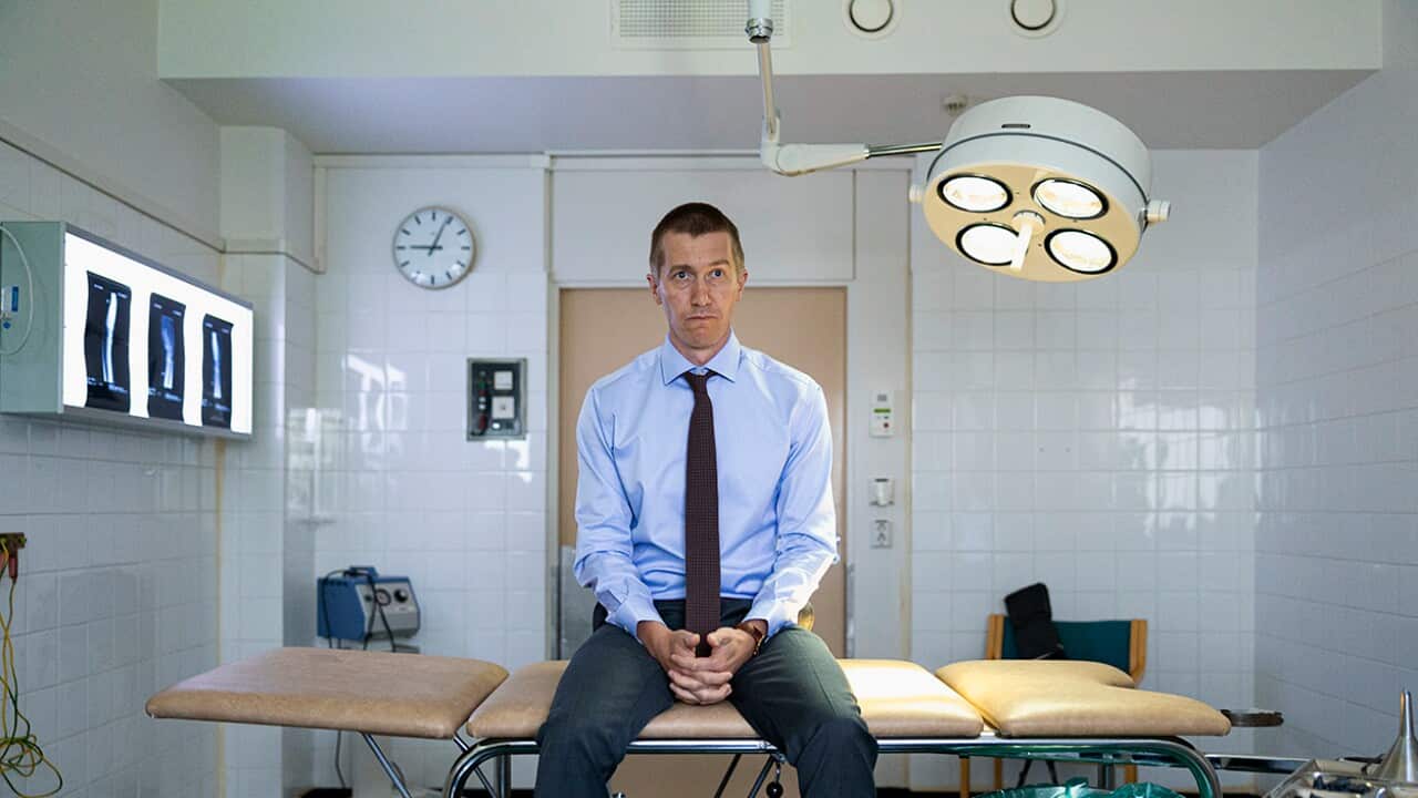 A man in a blue shirt and tie sits on an examination bench in a medical office, looking blankly ahead.