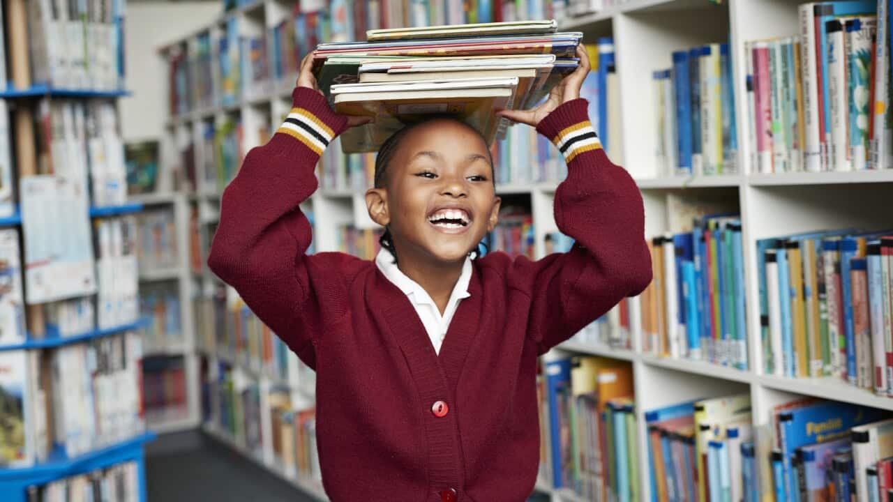 Msichana atabasamu akibeba vitabu juu ya kichwa ndani ya maktaba.