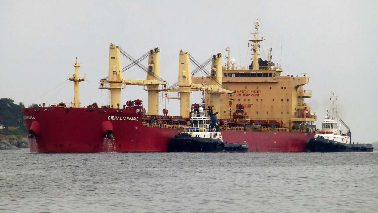 A bulk carrier with a red hull and two tug boats next to it.