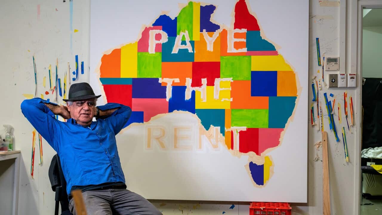 Richard bell reclines in a blue shirt and fedora in front of a painting, a multicoloured patchwork of australia with the words 'Pay the rent' superimposed on them.