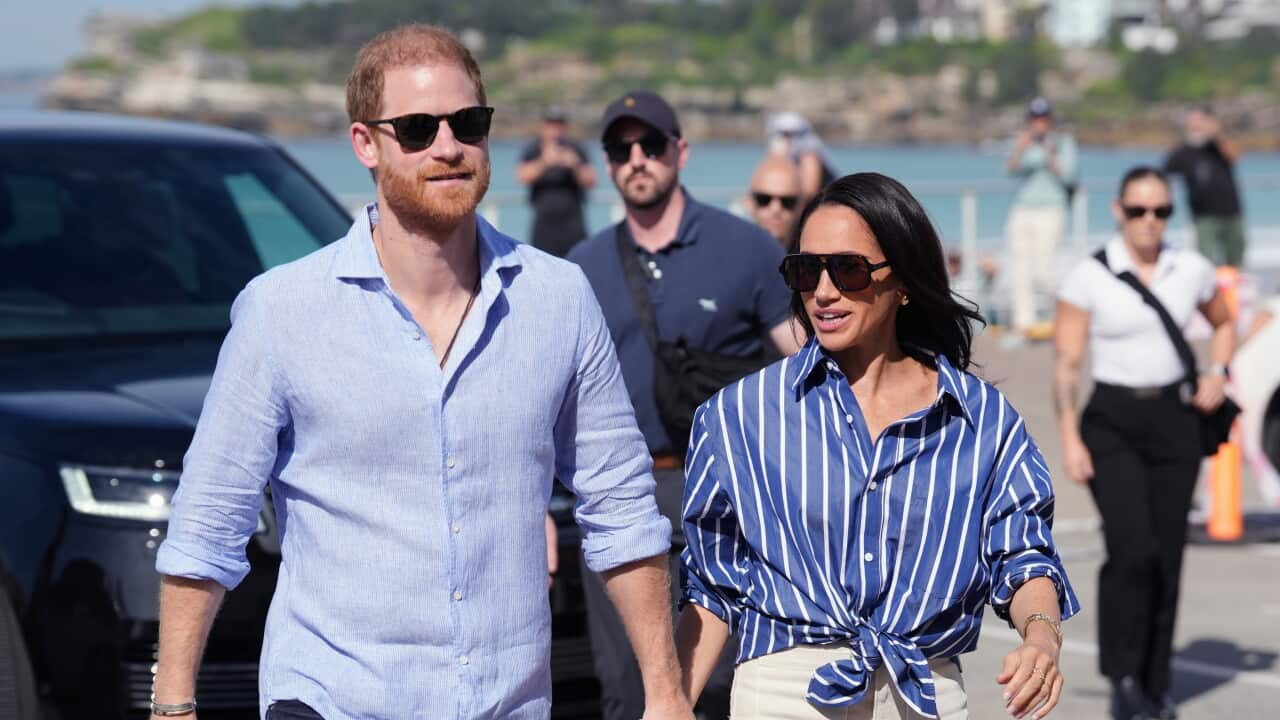 A man wearing a blue shirt and black sunglasses holds hands with a woman wearing a blue and white shirt as they walk