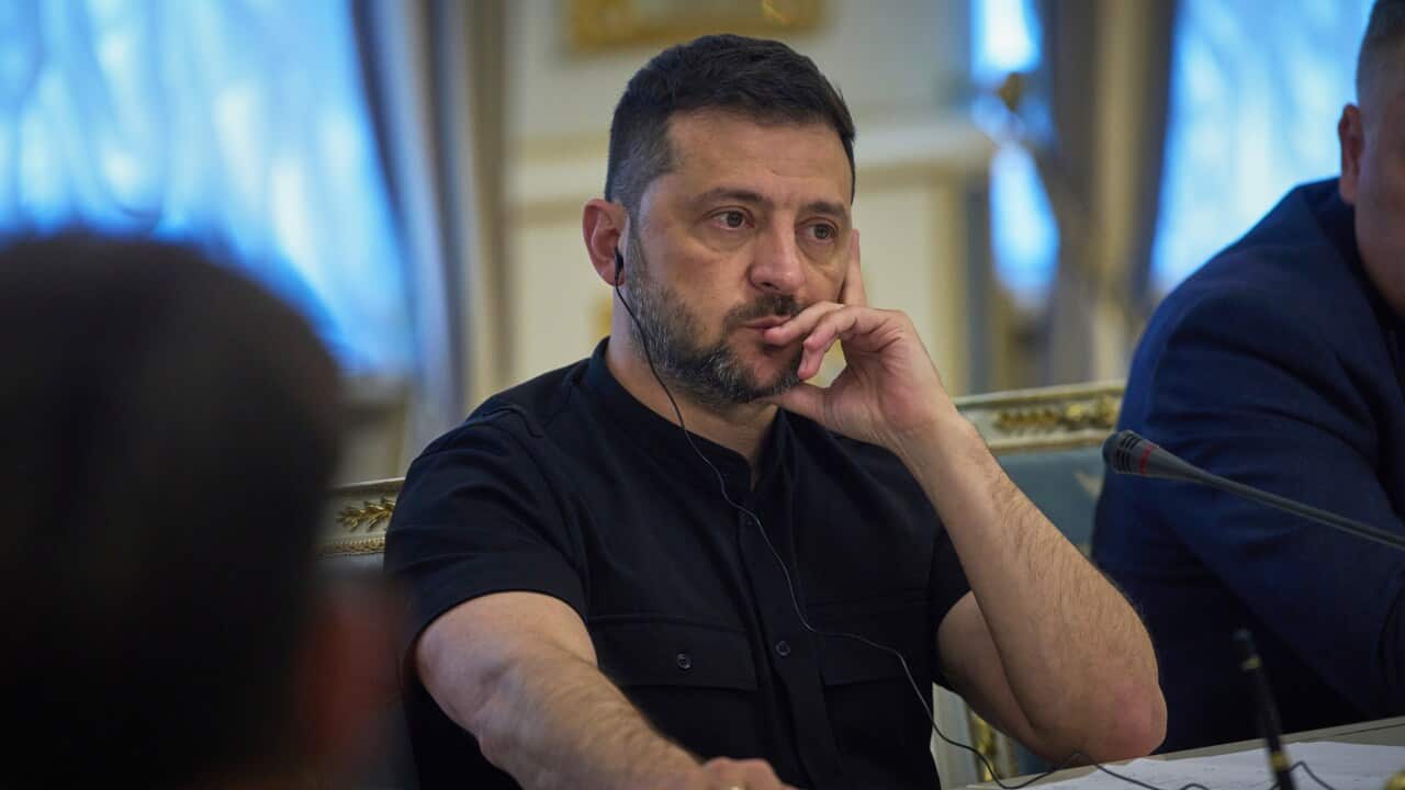 Volodymyr Zelensky, wearing a round, black T-shirt, sits with a serious expression, his hand on his face and the other on the table.