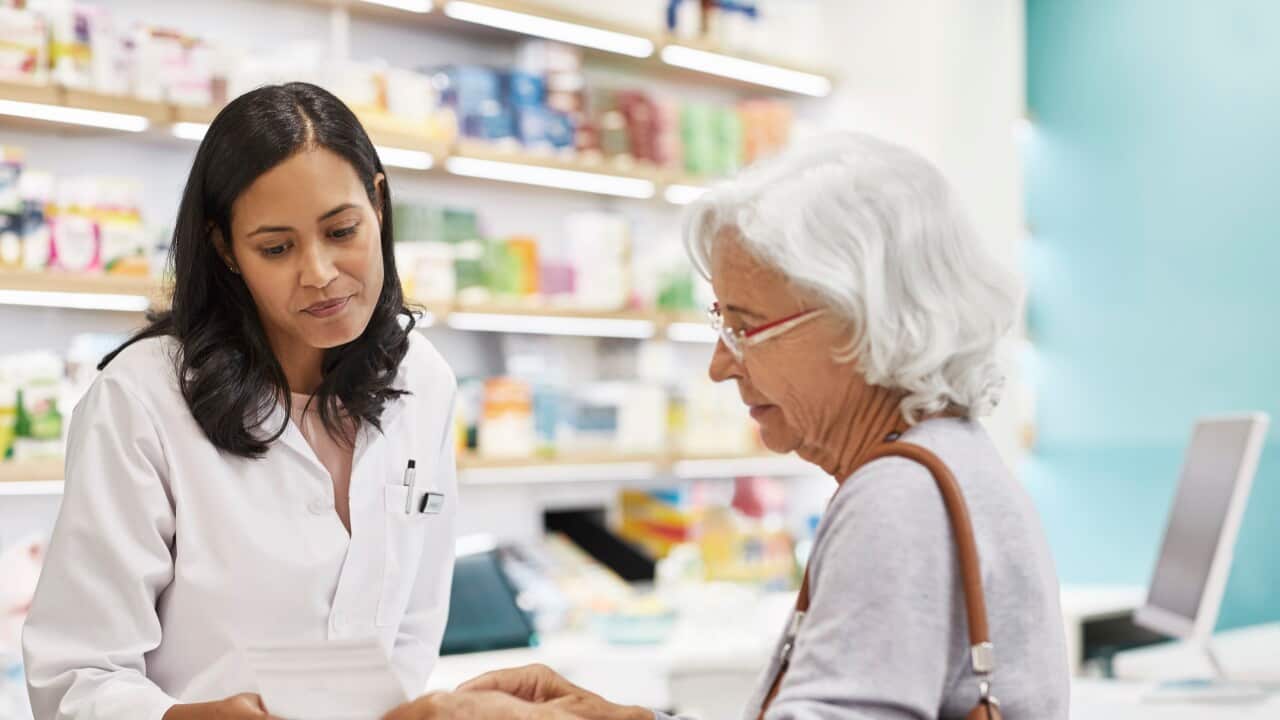 A pharmacist in a white coat reviews a prescription while consulting with an elderly woman at a pharmacy counter.