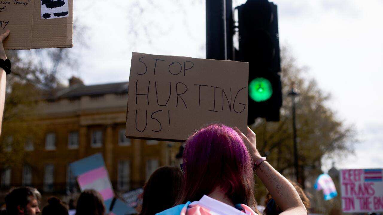A woman with a trans flag around her neck holds a sign that reads: "stop hurting us".