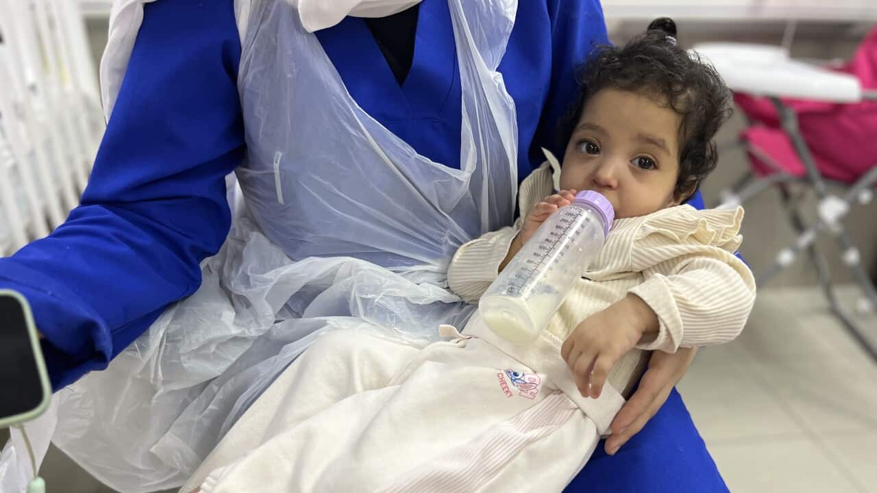 A baby is nursed from a bottle on the lap of a person wearing blue trousers