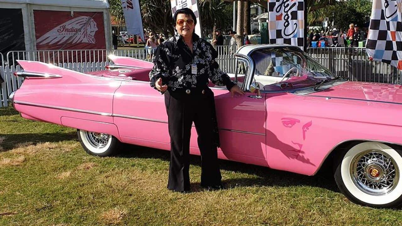 A woman dressed like Elvis standing outside next to a pink Cadillac.