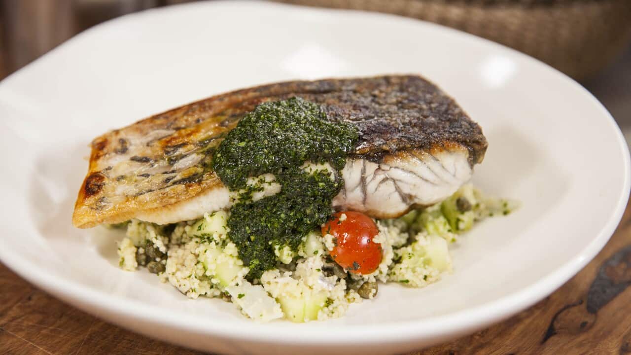 A fillet of fish with crispy skin sits on a bed of cousous on a white plate. A green sauce saouce has been drizzled over the fish and salad.