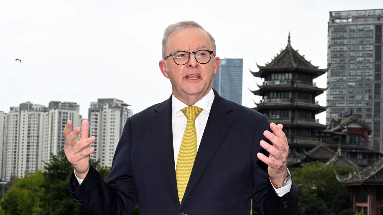 A man in a dark suit with yellow tie stands in front of a building with tiered pagoda roofs