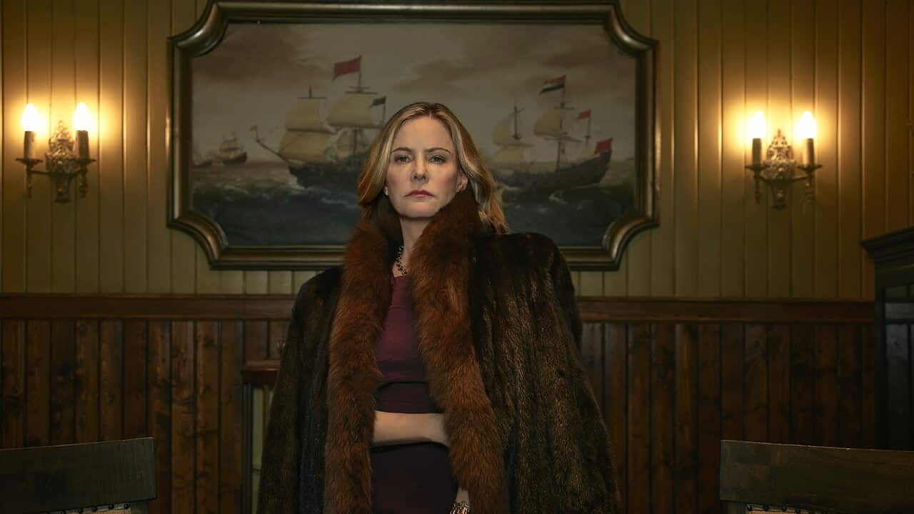 A woman in a dark fur coat stands in front of a wood-panelled wall in a dimly lit room.