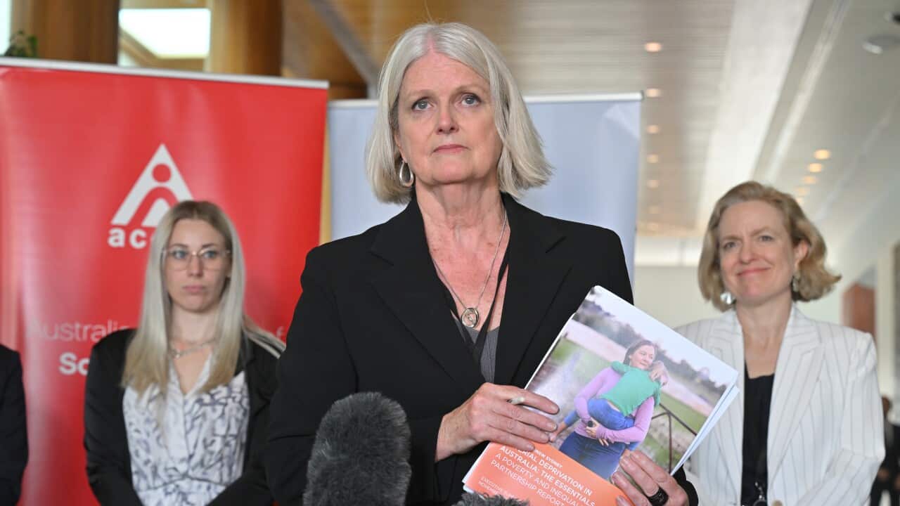 A woman stands in front of two microphones holding a report, with two women in the background to either side of her