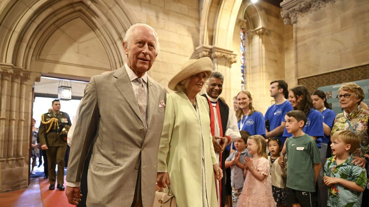 A man in a suit and a woman wearing a hat walk side by side as children and people stand by the side to greet them.