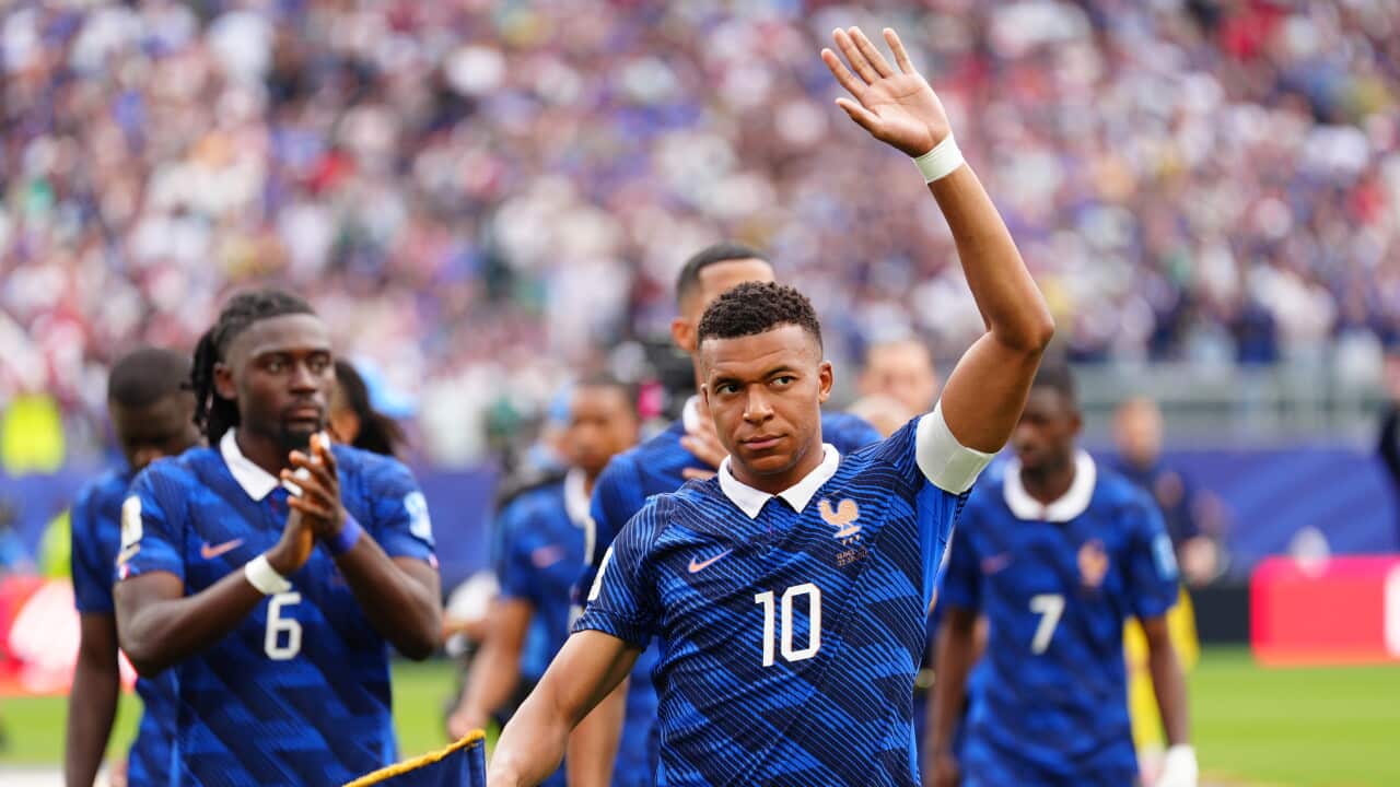 A footballer wearing a blue jersey waves to the crowd. His teammates stand behind him clapping as the crowd cheers.